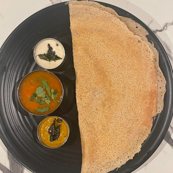 A large, crispy dosa with three small bowls of chutneys and sambar on a black plate.