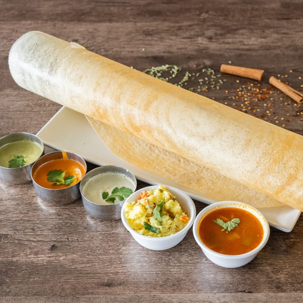 Rolled dosa on a white plate with five small bowls of chutneys and sambar on a wooden table.