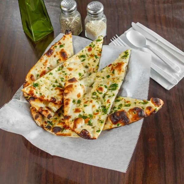 Garlic naan bread slices with green herbs in a basket, with salt, pepper, and cutlery.