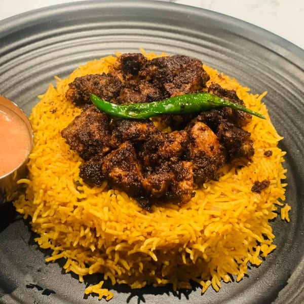 Indian chicken biryani with yellow rice, dark chicken pieces, a green chili, and a side of sauce.