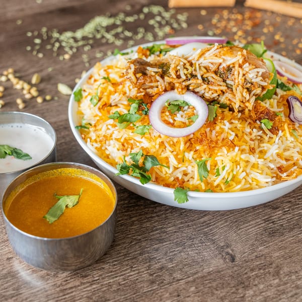 Indian chicken biryani with rice, onions, cilantro, and two side sauces on a wooden table.