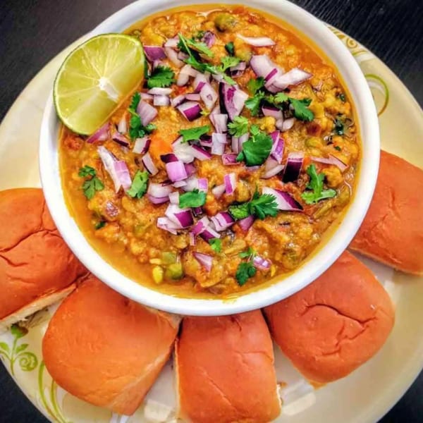 A bowl of Cumin N Eat's Indian pav bhaji, garnished with lime, red onions, and cilantro, served with pav.