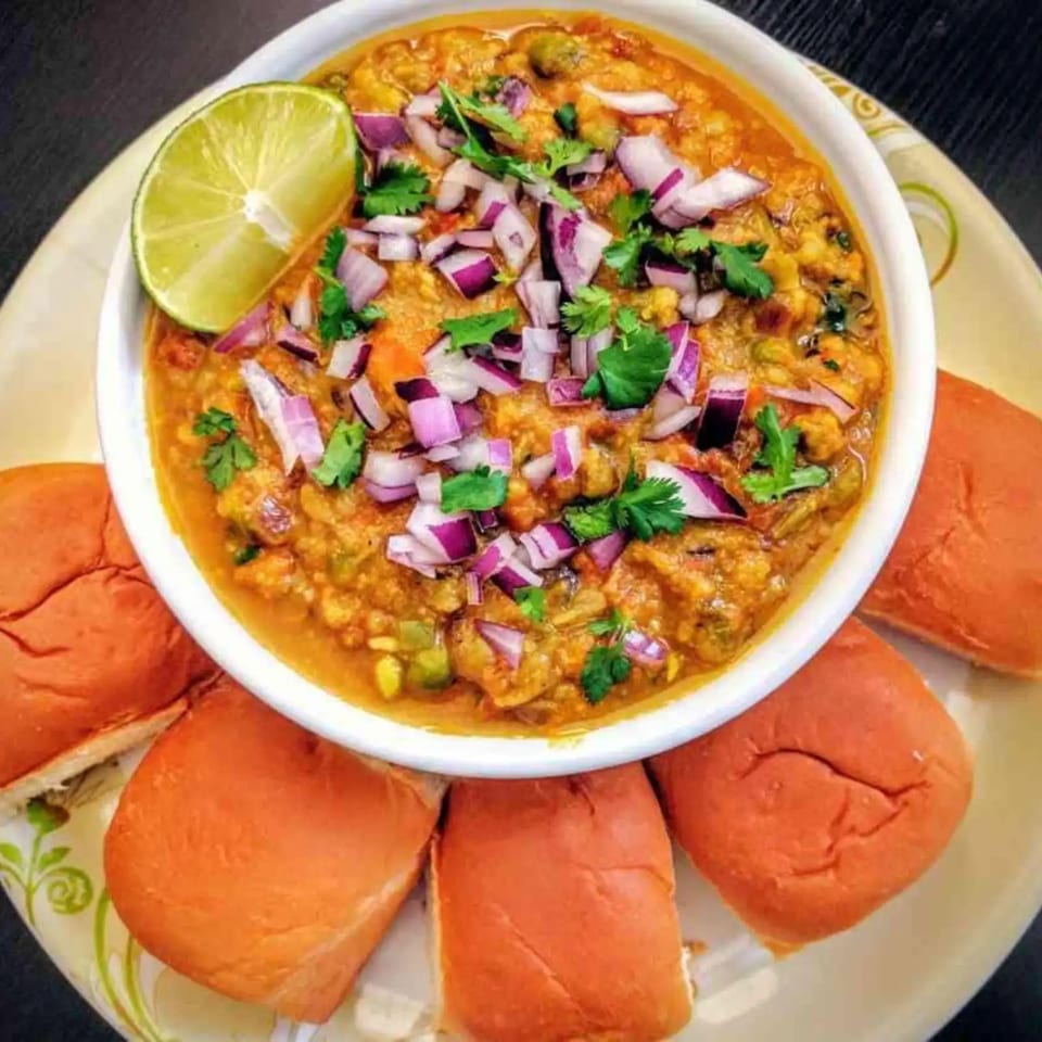 A bowl of Cumin N Eat's Indian pav bhaji, garnished with lime, red onions, and cilantro, served with pav.